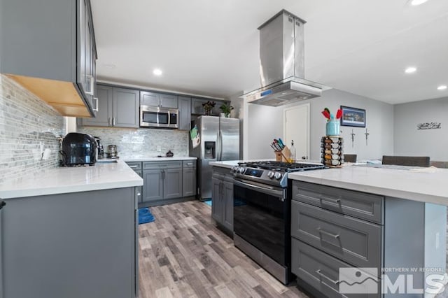 kitchen with appliances with stainless steel finishes, gray cabinets, wood-type flooring, tasteful backsplash, and island exhaust hood