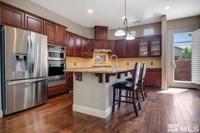 kitchen with light stone counters, dark hardwood / wood-style flooring, stainless steel appliances, hanging light fixtures, and a kitchen island with sink