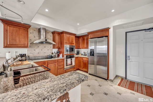 kitchen with light stone counters, backsplash, wall chimney exhaust hood, appliances with stainless steel finishes, and sink