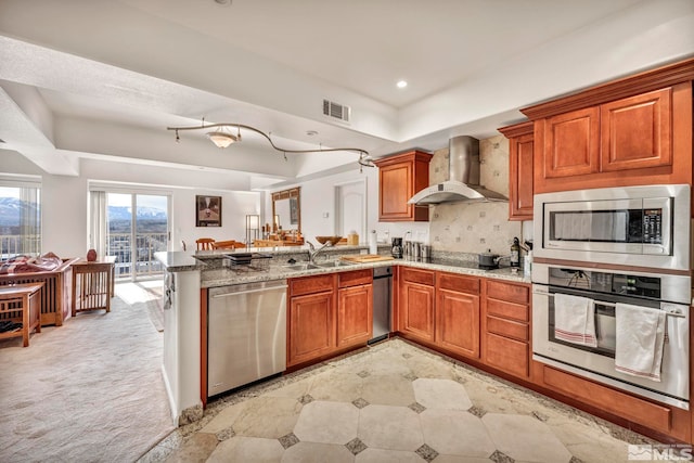 kitchen with kitchen peninsula, tasteful backsplash, light stone counters, wall chimney exhaust hood, and appliances with stainless steel finishes