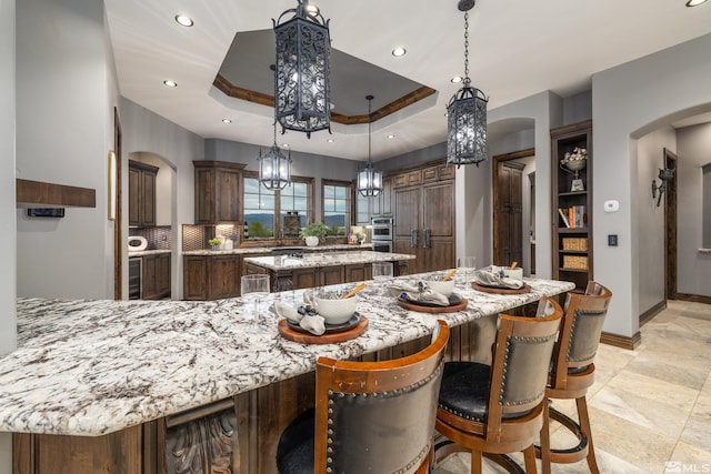 kitchen with a spacious island, light stone counters, decorative light fixtures, light tile floors, and a raised ceiling