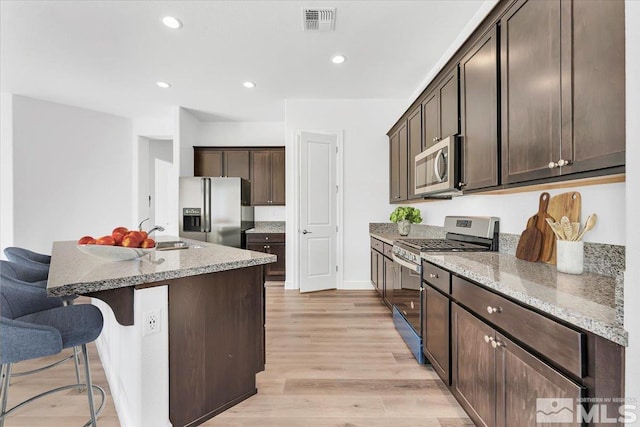 kitchen featuring dark brown cabinets, a kitchen island with sink, light hardwood / wood-style flooring, appliances with stainless steel finishes, and light stone countertops