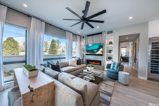 living room featuring a large fireplace, built in features, ceiling fan, and light hardwood / wood-style flooring