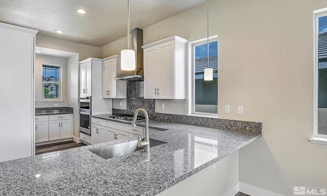 kitchen with white cabinetry, sink, hanging light fixtures, wall chimney range hood, and kitchen peninsula