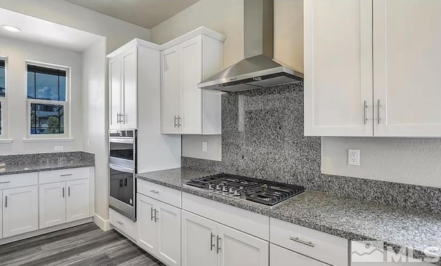 kitchen featuring appliances with stainless steel finishes, dark stone counters, dark wood-type flooring, wall chimney range hood, and white cabinets