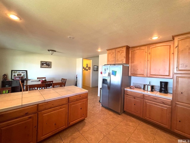 kitchen with a textured ceiling, light tile flooring, stainless steel refrigerator with ice dispenser, and tile counters