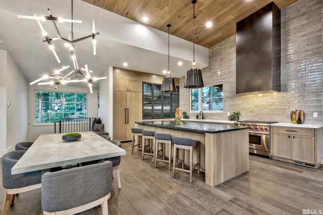 kitchen with light brown cabinetry, wall chimney range hood, hanging light fixtures, and stainless steel stove