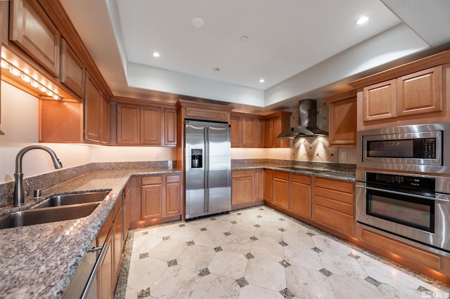 kitchen with sink, a tray ceiling, dark stone counters, wall chimney range hood, and stainless steel appliances