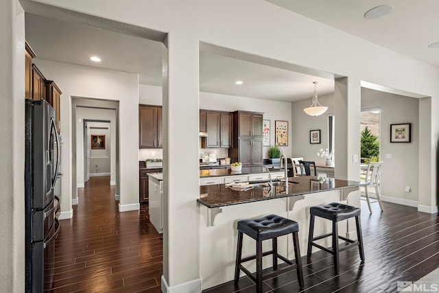 kitchen with hanging light fixtures, dark hardwood / wood-style flooring, stainless steel fridge with ice dispenser, dark stone countertops, and a kitchen island with sink