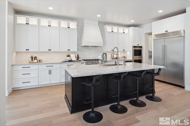 kitchen with white cabinets, a center island with sink, premium range hood, and stainless steel appliances