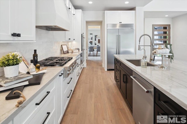 kitchen with light stone counters, custom exhaust hood, stainless steel appliances, sink, and white cabinetry