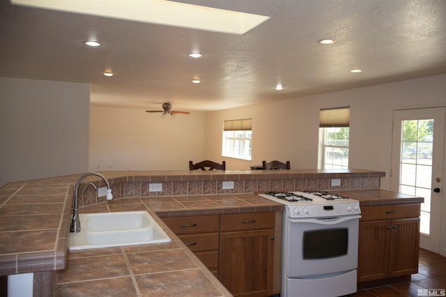 kitchen with white gas range oven, ceiling fan, sink, dark tile patterned flooring, and tile counters