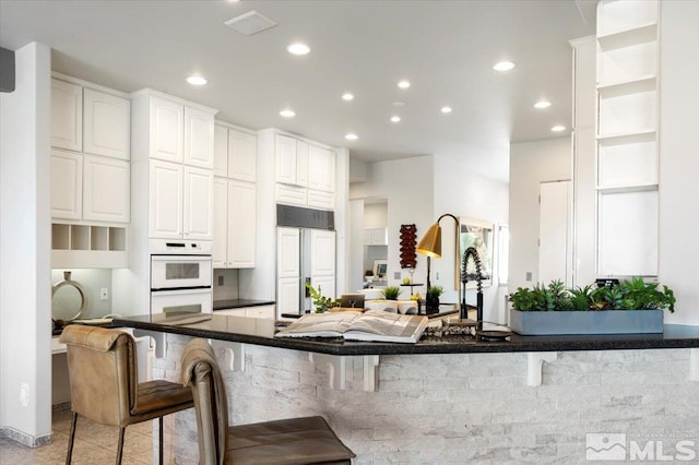 kitchen with paneled fridge, dark countertops, double oven, a kitchen bar, and white cabinetry