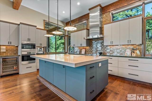 kitchen featuring pendant lighting, white cabinetry, beverage cooler, a kitchen island with sink, and wall chimney exhaust hood