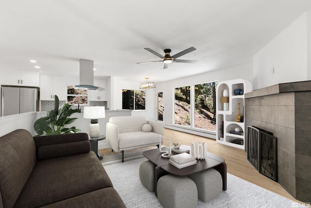 living room with baseboard heating, ceiling fan, a tiled fireplace, and light wood-type flooring