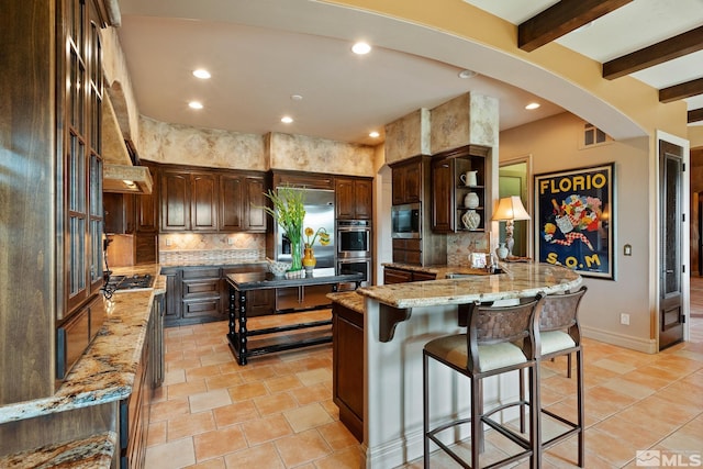 kitchen with built in appliances, beamed ceiling, light stone counters, dark brown cabinets, and backsplash