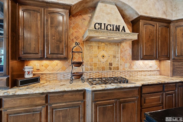 kitchen featuring stainless steel gas cooktop, premium range hood, light stone countertops, and backsplash