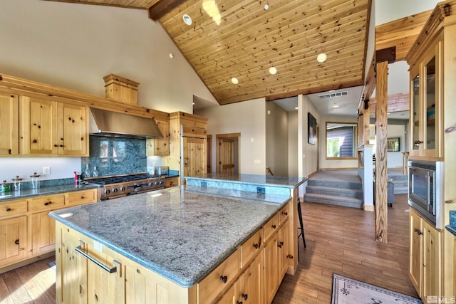 kitchen featuring appliances with stainless steel finishes, a kitchen island, dark stone counters, light brown cabinets, and wall chimney exhaust hood