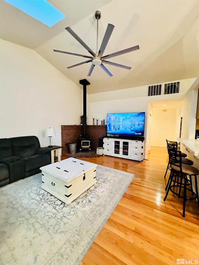 living room with ceiling fan, a wood stove, light hardwood / wood-style flooring, and lofted ceiling with skylight