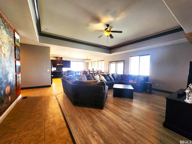 living area featuring baseboards, ornamental molding, a tray ceiling, light wood-type flooring, and ceiling fan with notable chandelier