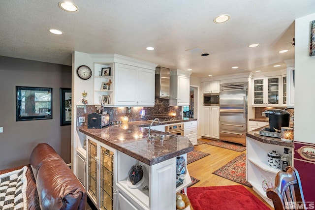 kitchen with built in appliances, open shelves, wall chimney range hood, and white cabinets