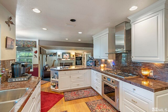 kitchen with light wood-type flooring, stainless steel appliances, decorative backsplash, wall chimney range hood, and kitchen peninsula