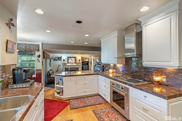 kitchen featuring stainless steel appliances, white cabinetry, wall chimney range hood, and a peninsula