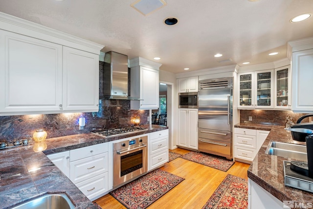 kitchen with dark countertops, white cabinetry, wall chimney exhaust hood, and built in appliances