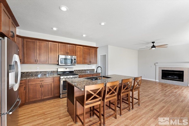 kitchen with light wood-type flooring, appliances with stainless steel finishes, a tiled fireplace, ceiling fan, and sink