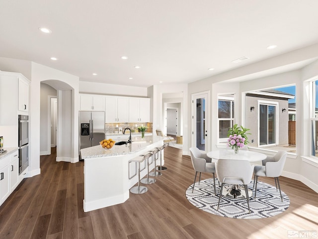 kitchen featuring arched walkways, stainless steel appliances, an island with sink, and white cabinets