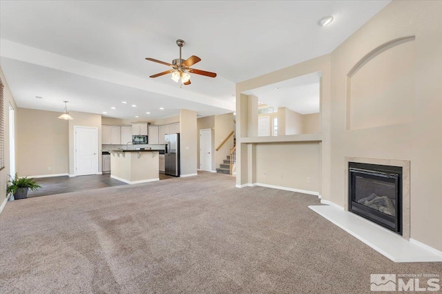 unfurnished living room featuring dark colored carpet, a glass covered fireplace, ceiling fan, baseboards, and stairs