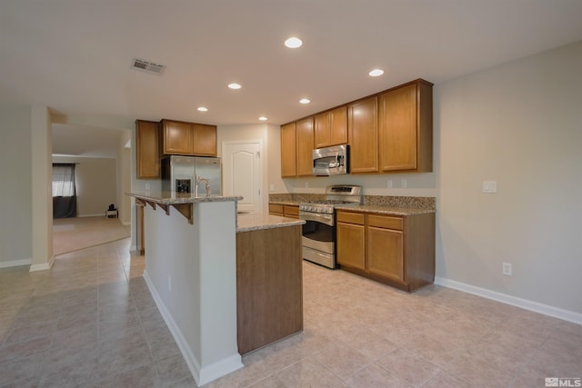 kitchen with appliances with stainless steel finishes, light tile patterned flooring, a kitchen island with sink, and light stone counters