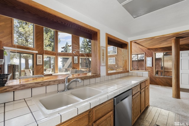 kitchen featuring sink, stainless steel dishwasher, wooden walls, tile counters, and light colored carpet