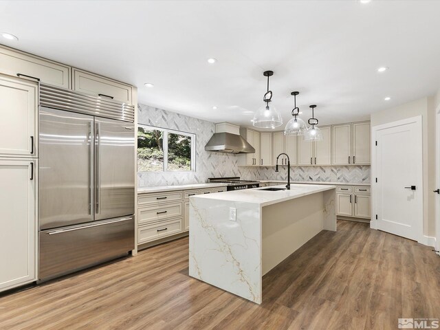 kitchen with wall chimney exhaust hood, built in fridge, a kitchen island with sink, and hardwood / wood-style floors