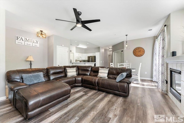 living room with a ceiling fan, wood finished floors, baseboards, a tile fireplace, and vaulted ceiling