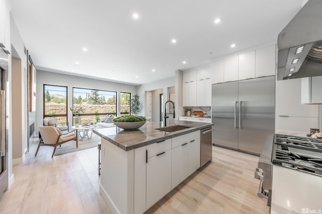 kitchen with wall chimney range hood, an island with sink, appliances with stainless steel finishes, light hardwood / wood-style floors, and sink