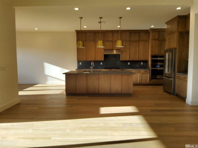 kitchen with hanging light fixtures, decorative backsplash, light wood-type flooring, an island with sink, and stainless steel appliances