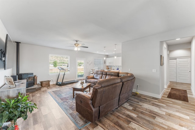 living room with a wood stove, light wood finished floors, ceiling fan, and baseboards