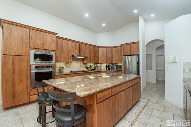 kitchen with brown cabinetry, appliances with stainless steel finishes, light stone counters, a center island, and high vaulted ceiling