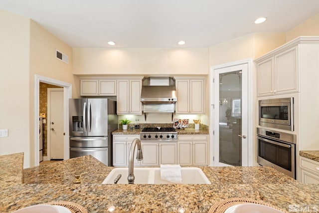 kitchen featuring appliances with stainless steel finishes, light stone countertops, sink, wall chimney range hood, and cream cabinetry