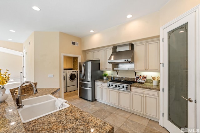 kitchen featuring stainless steel appliances, washing machine and clothes dryer, sink, cream cabinets, and wall chimney range hood