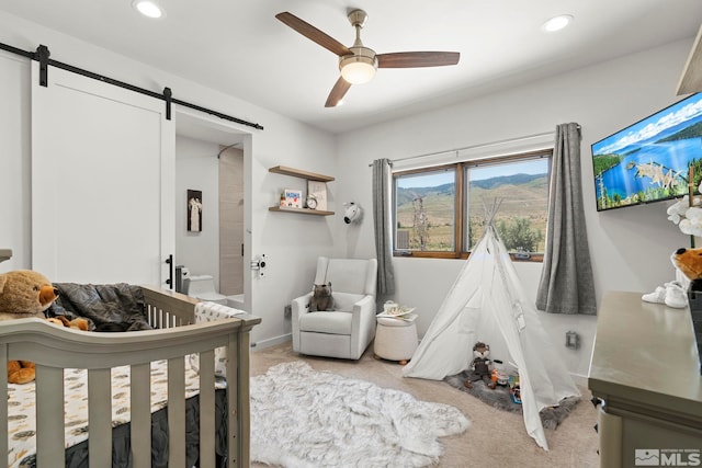 bedroom featuring a barn door, a nursery area, light colored carpet, and ceiling fan