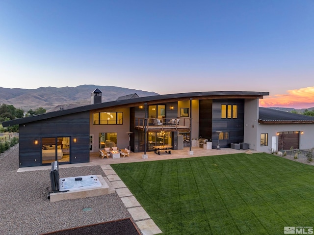 back house at dusk featuring a hot tub, a mountain view, a lawn, and a patio area