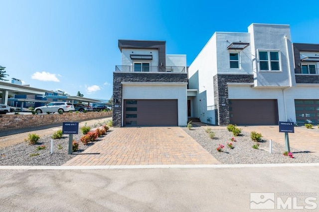 modern home featuring decorative driveway, stucco siding, a balcony, a garage, and stone siding