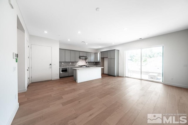 kitchen featuring tasteful backsplash, light wood-style flooring, gray cabinetry, under cabinet range hood, and stainless steel oven