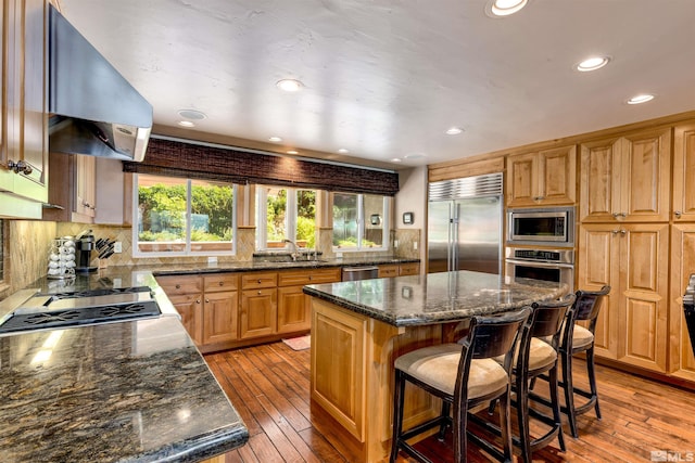 kitchen with a breakfast bar, a kitchen island, wall chimney range hood, built in appliances, and light wood-type flooring