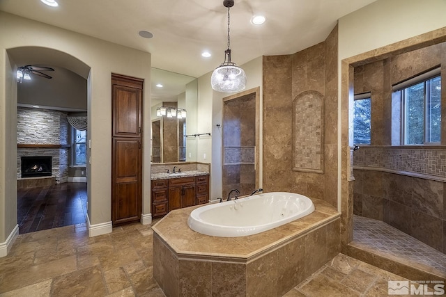 bathroom with stone tile floors, vanity, a warm lit fireplace, baseboards, and a bath