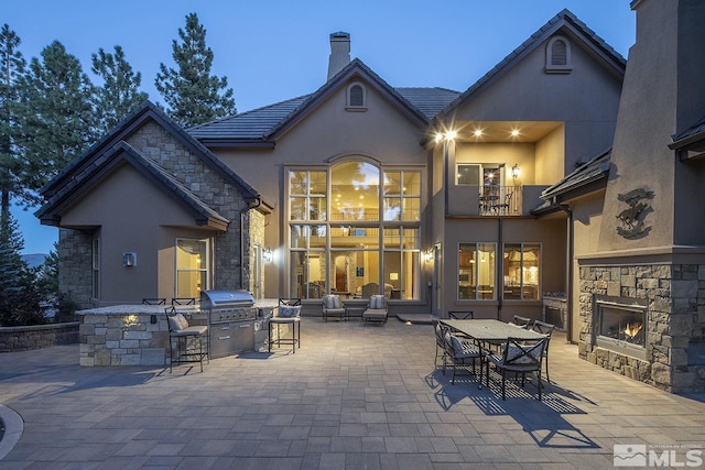 back of house with an outdoor stone fireplace, a chimney, a patio area, a balcony, and exterior kitchen