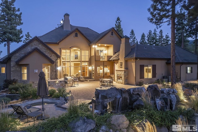 back of house with a balcony, stone siding, stucco siding, a chimney, and a patio area