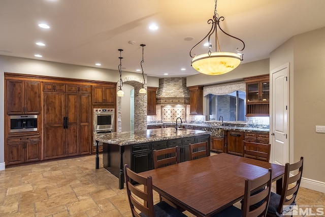 kitchen featuring an island with sink, premium range hood, stone tile flooring, and recessed lighting